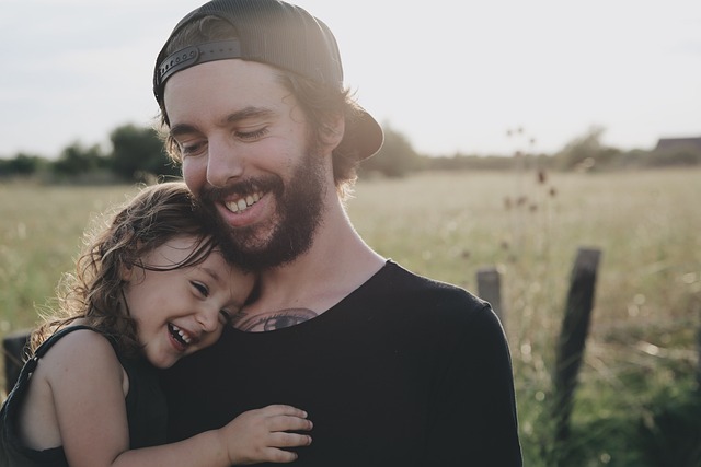 moment tendre entre un père et sa fille. Ils sont tous les deux souriants et se tiennent dans les bras l’un de l’autre, dans un cadre champêtre baigné de lumière naturelle. L’homme porte une casquette à l’envers et un t-shirt noir, tandis que la petite fille, hilare, semble pleinement heureuse et en sécurité. L’ambiance est chaleureuse, simple et empreinte de complicité.