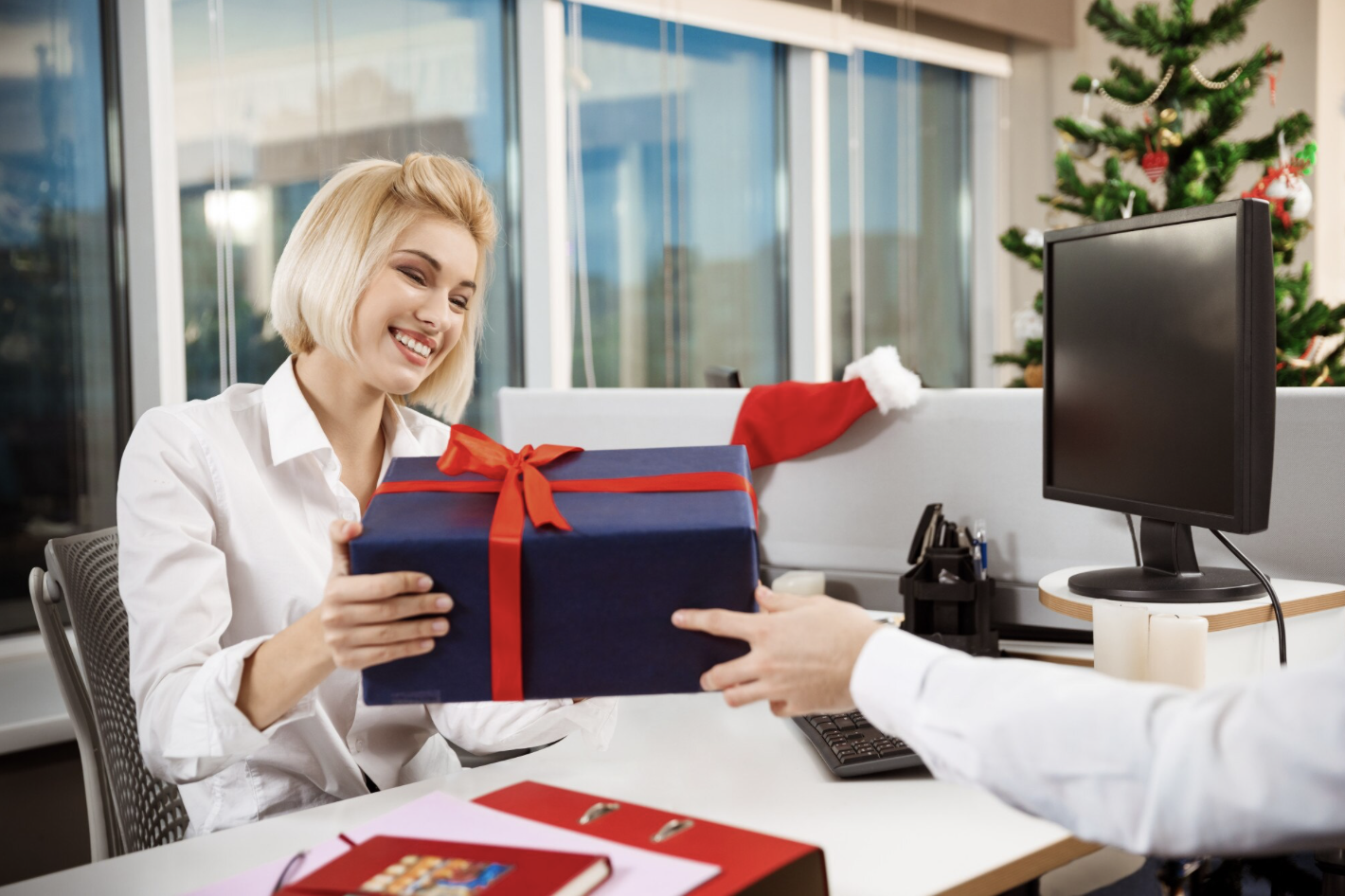 Cette image montre une scène festive au bureau. Une femme souriante reçoit un cadeau emballé d’un ruban rouge, probablement offert par un collègue. En arrière-plan, on aperçoit un sapin de Noël décoré et un bonnet de Père Noël posé sur une cloison de bureau, ce qui suggère un échange de cadeaux dans le cadre des fêtes de fin d’année en entreprise. L’ambiance est chaleureuse et conviviale.