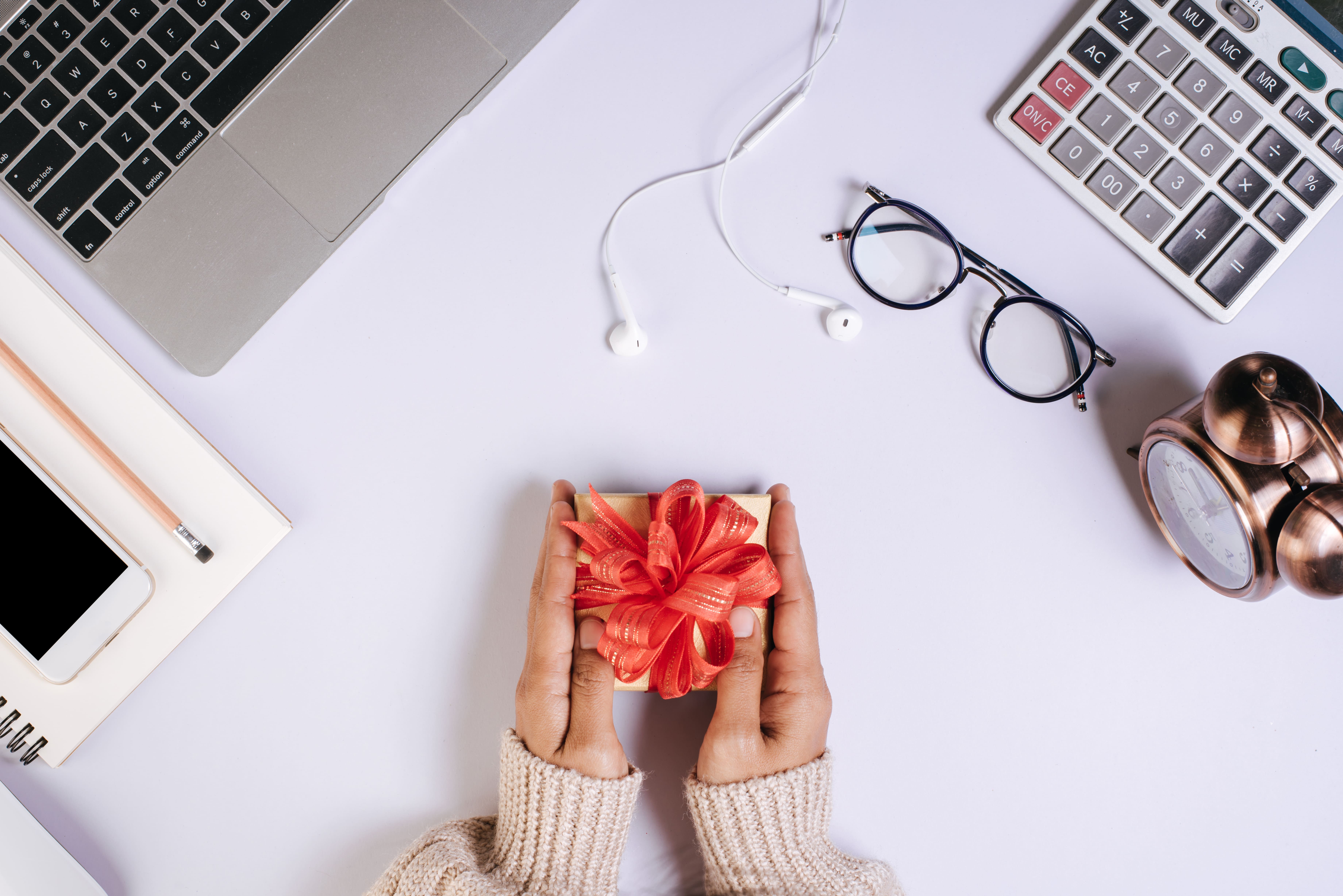 Cette image montre une vue de dessus d’un bureau sur lequel sont disposés un ordinateur portable, un téléphone, une paire de lunettes, une calculatrice, une horloge et des écouteurs. Au centre, une personne tient un petit coffret cadeau doré décoré d’un nœud rouge. L’ensemble évoque une surprise ou une attention offerte dans un cadre professionnel, symbolisant la reconnaissance ou un moment spécial au travail.