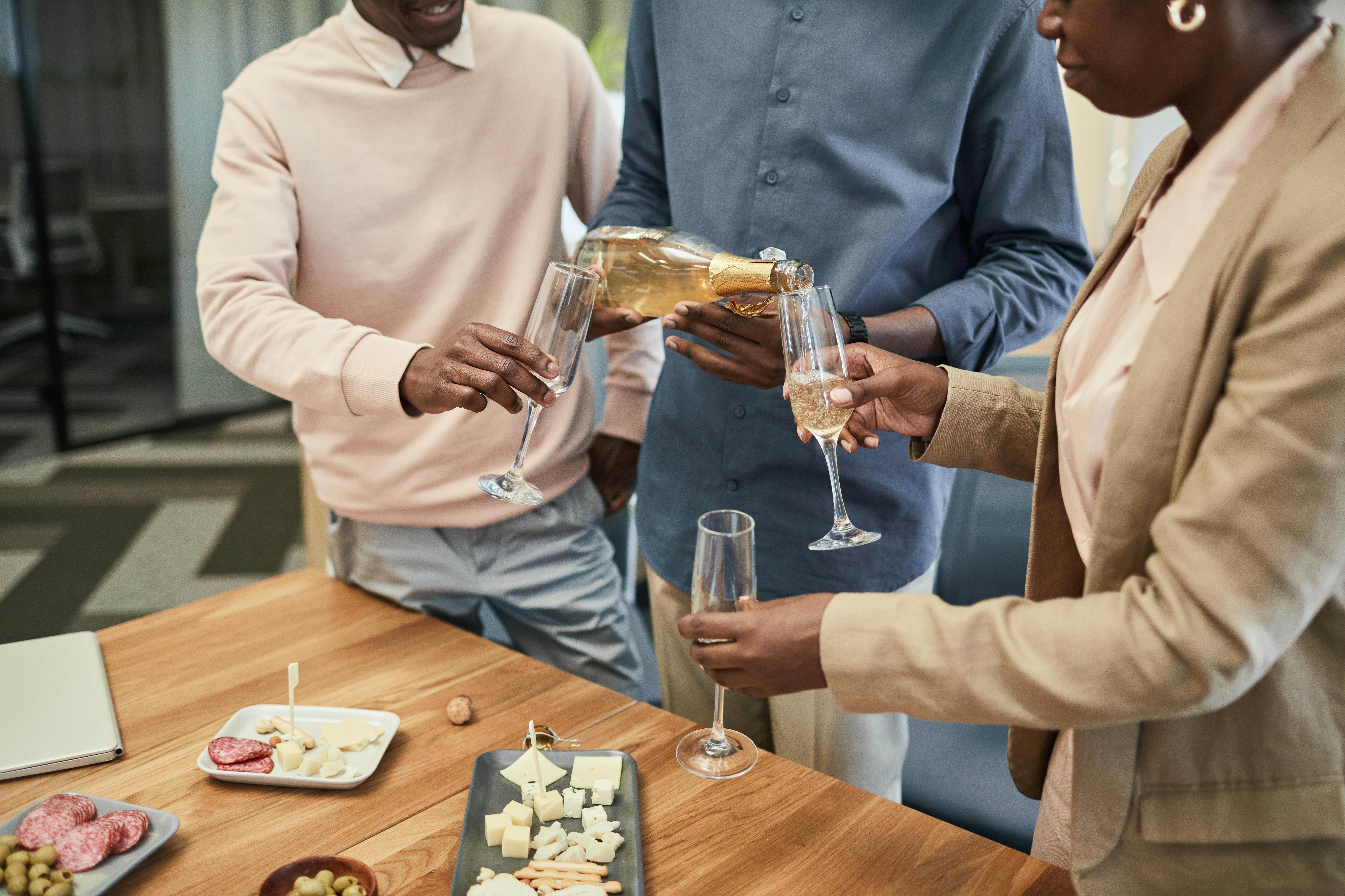 Cette image montre un moment convivial entre collègues autour d’un apéritif. Trois personnes échangent des verres de champagne, tandis que l’une d’elles sert la boisson. Sur la table, on aperçoit des plateaux de fromages, de charcuterie et des olives. L’ambiance est détendue et chaleureuse, suggérant une célébration informelle au bureau, peut-être à l’occasion d’un succès d’équipe, d’un départ ou d’un événement d’entreprise.