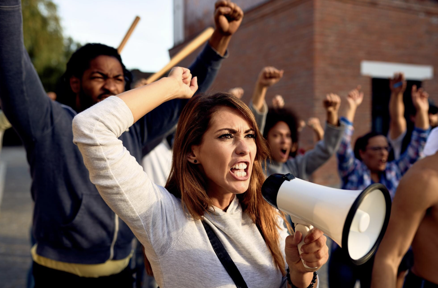 Cette image montre une scène de manifestation. Au premier plan, une femme tient un mégaphone et s’exprime avec intensité, poing levé, symbole de protestation. Derrière elle, un groupe de personnes manifeste également, toutes avec le poing en l’air, exprimant colère, revendication ou mobilisation collective. L’ambiance est tendue et déterminée, typique d’un mouvement de revendication sociale ou syndicale.