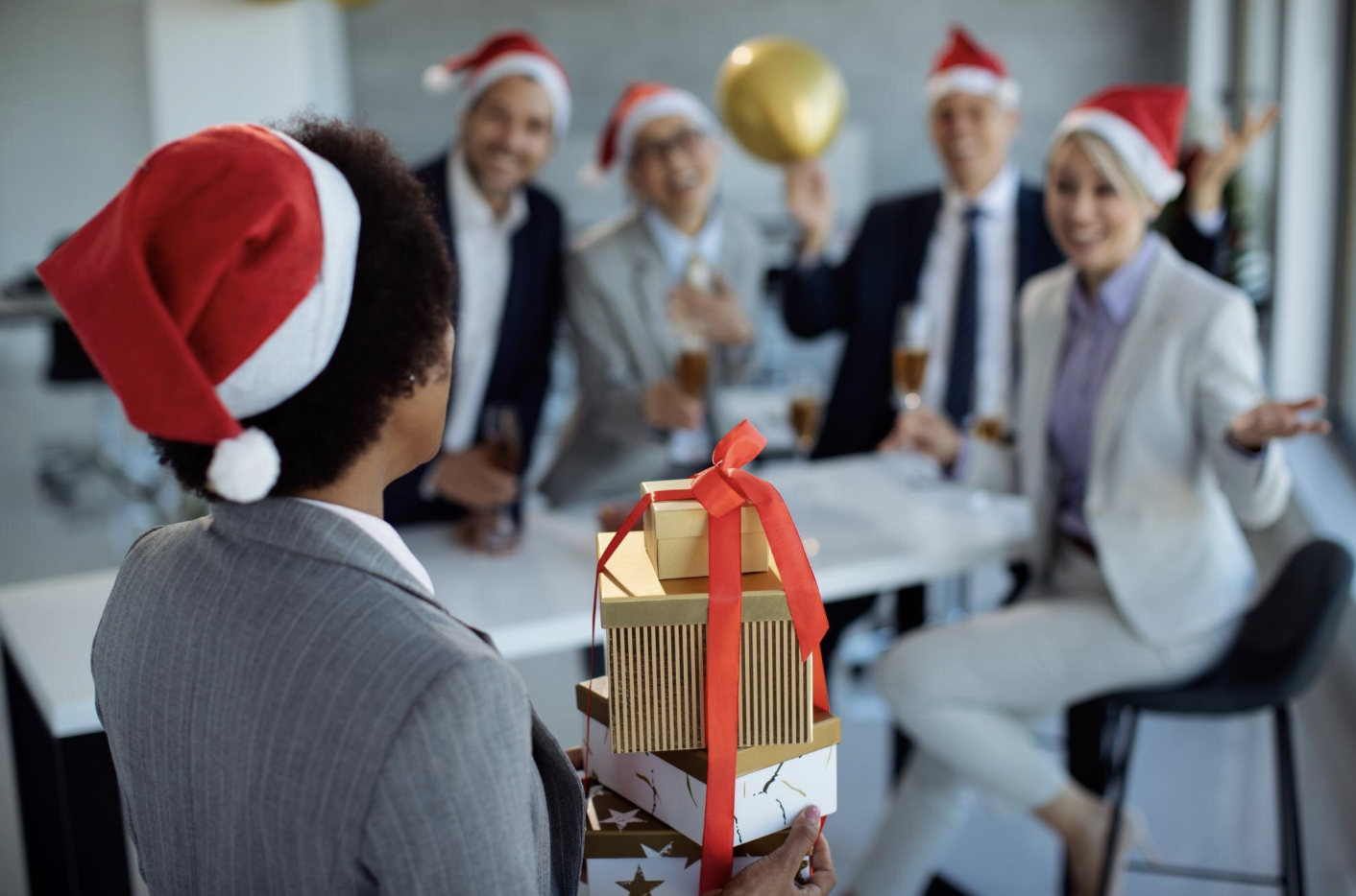 Cette image montre une scène festive en entreprise, probablement lors d’une célébration de Noël. Une personne en costume, de dos, portant un bonnet de Noël, tient une pile de cadeaux joliment emballés avec un ruban rouge. En face, un groupe de collègues souriants, également coiffés de bonnets de Noël, semblent l’accueillir avec enthousiasme, verre à la main. L’ambiance est conviviale et joyeuse, illustrant un moment de partage et de reconnaissance en milieu professionnel.