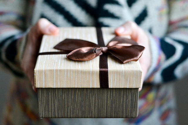 Cette image montre une personne tenant un coffret cadeau entre ses mains. La boîte est élégante, avec un couvercle beige à fines rayures, décoré d’un ruban marron satiné noué en nœud. Le bas de la boîte est d’un ton plus foncé, contrastant avec le couvercle. L’arrière-plan est flou, mettant en valeur le cadeau, ce qui évoque un geste attentionné, un moment de partage ou une surprise à offrir.
